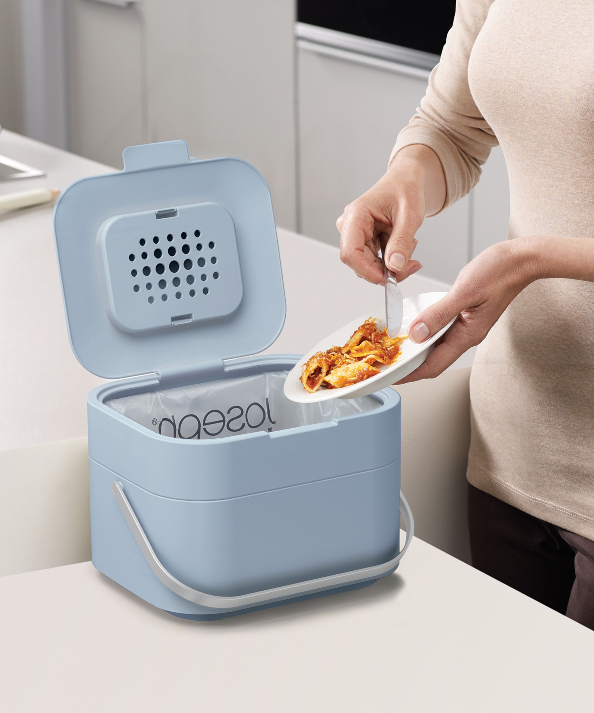A compact blue food waste caddy with a handle and vented lid, placed on a kitchen counter as food scraps are scraped in from a plate.
