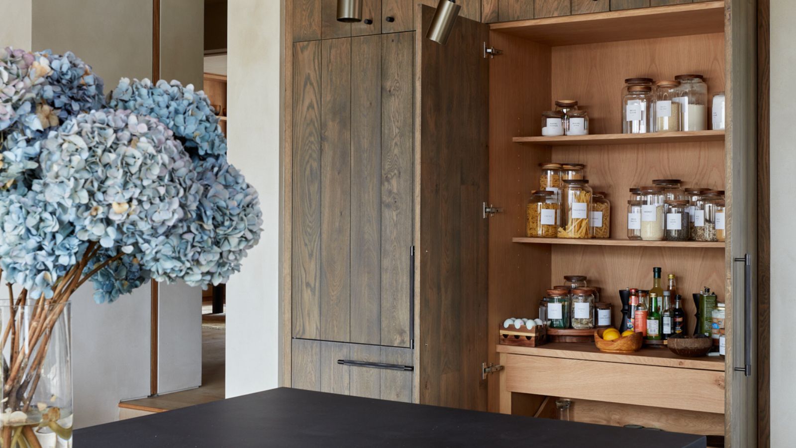 Minimalist pantry cabinet with warm wood interiors, soft oak shelving, and neatly labelled jars, blending seamlessly into a calm, modern kitchen.