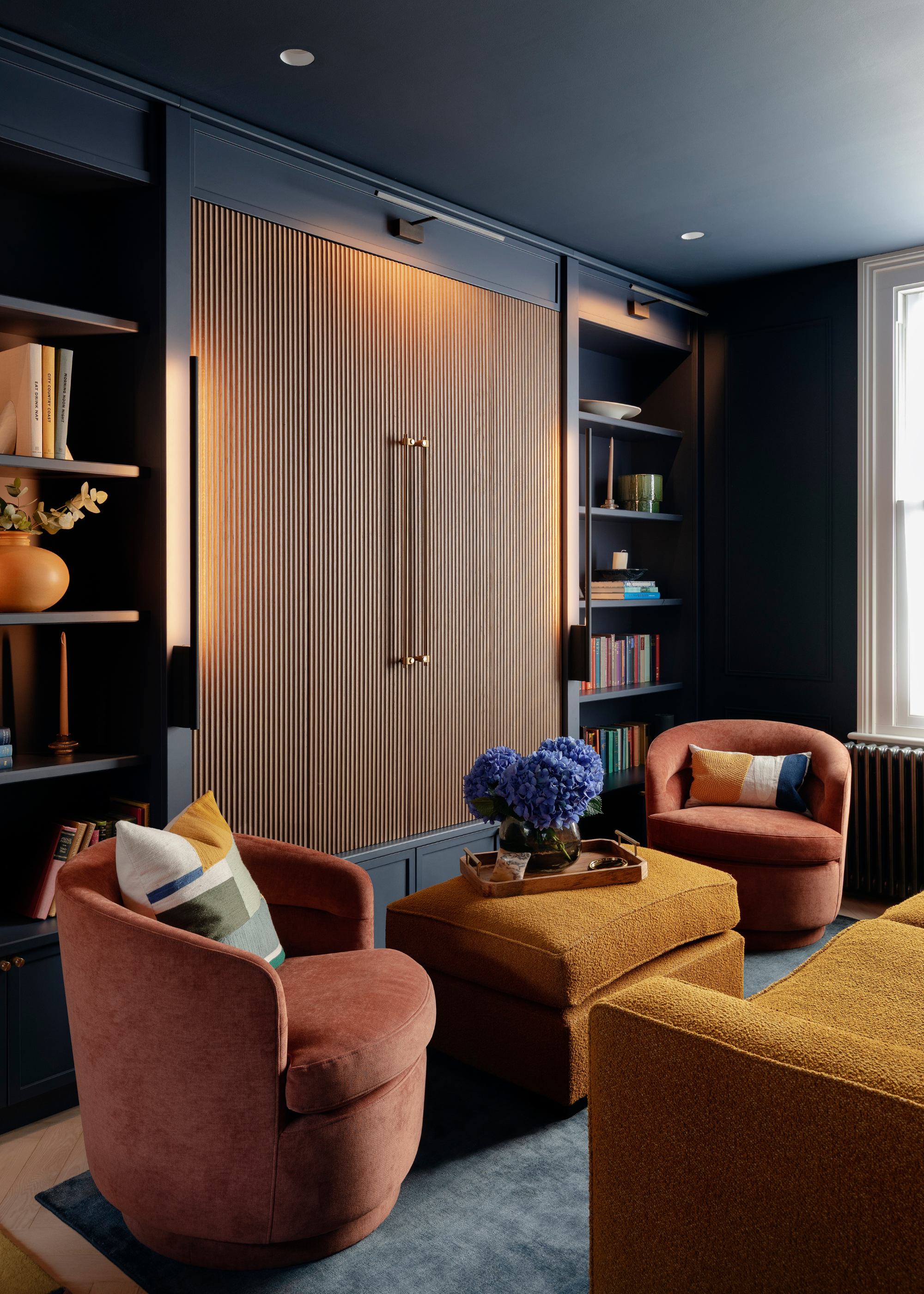 A navy entertainment room with a pair of tan swivel armchairs with patterned cushions, a storage pouf with a tray and a vase of blue hydrangeas beside a TV unit with wood panelled doors in between two storage units with shelves of books, tapered candles, and ceramics
