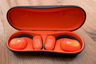 Bright orange Oladance open ear headphones nestled in the case of matching colour which connects to a charger, on a wooden surface