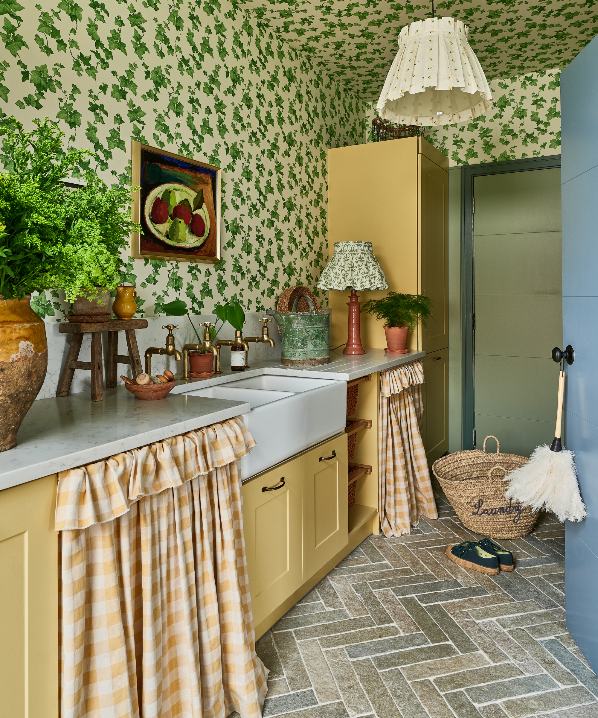 utility room with green floral wallpaper, yellow cabinets, limestone tiled floor, white butler sink, fabric lightshade, plants, curtains covering shelving below sink and wicker laundry basket on floor