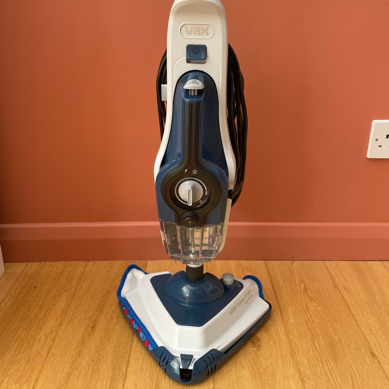 VAX Total Home Steam Cleaner sitting on wooden floor in front of a pink painted wall