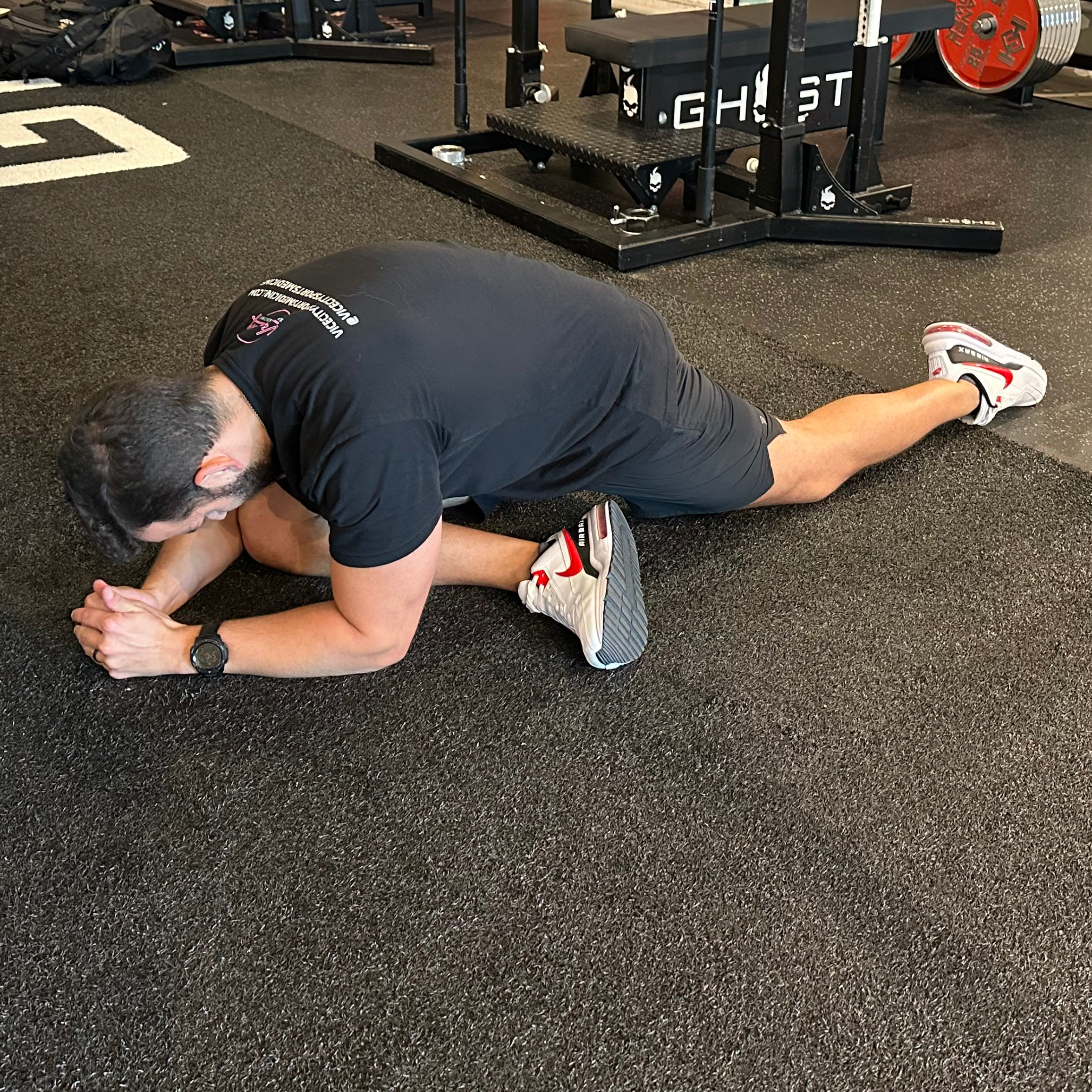 An employee at Vice City Sports Medicine performs a pigeon pose in a gym setting. They are on the floor, with their right shin horizontal to their body, forearms on the floor, torso bent forward and left leg behind them. 