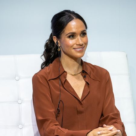 Meghan Markle wears her dark hair in a ponytail and wears a brown shirt and a gold necklace