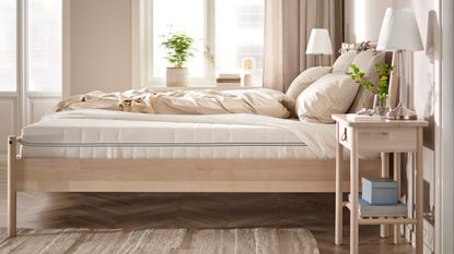 IKEA Abrotten mattress on a wooden bedframe in a neutral bedroom with wooden flooring, a brown rug, white bedside table with two white matching lamps, and a large window behind with a green pot plant.