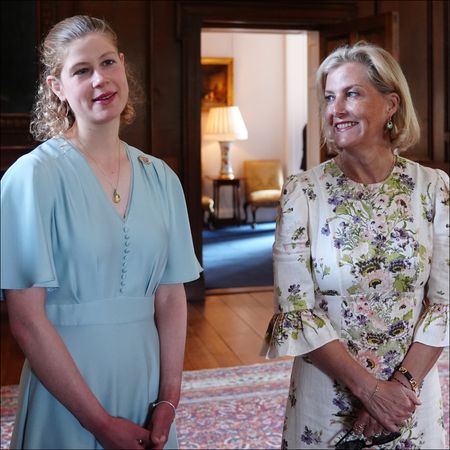 Lady Louise has blonde curly hair and is wearing a light blue dress while her mom Duchess Sophie has a blonde bob and is wearing a floral dress