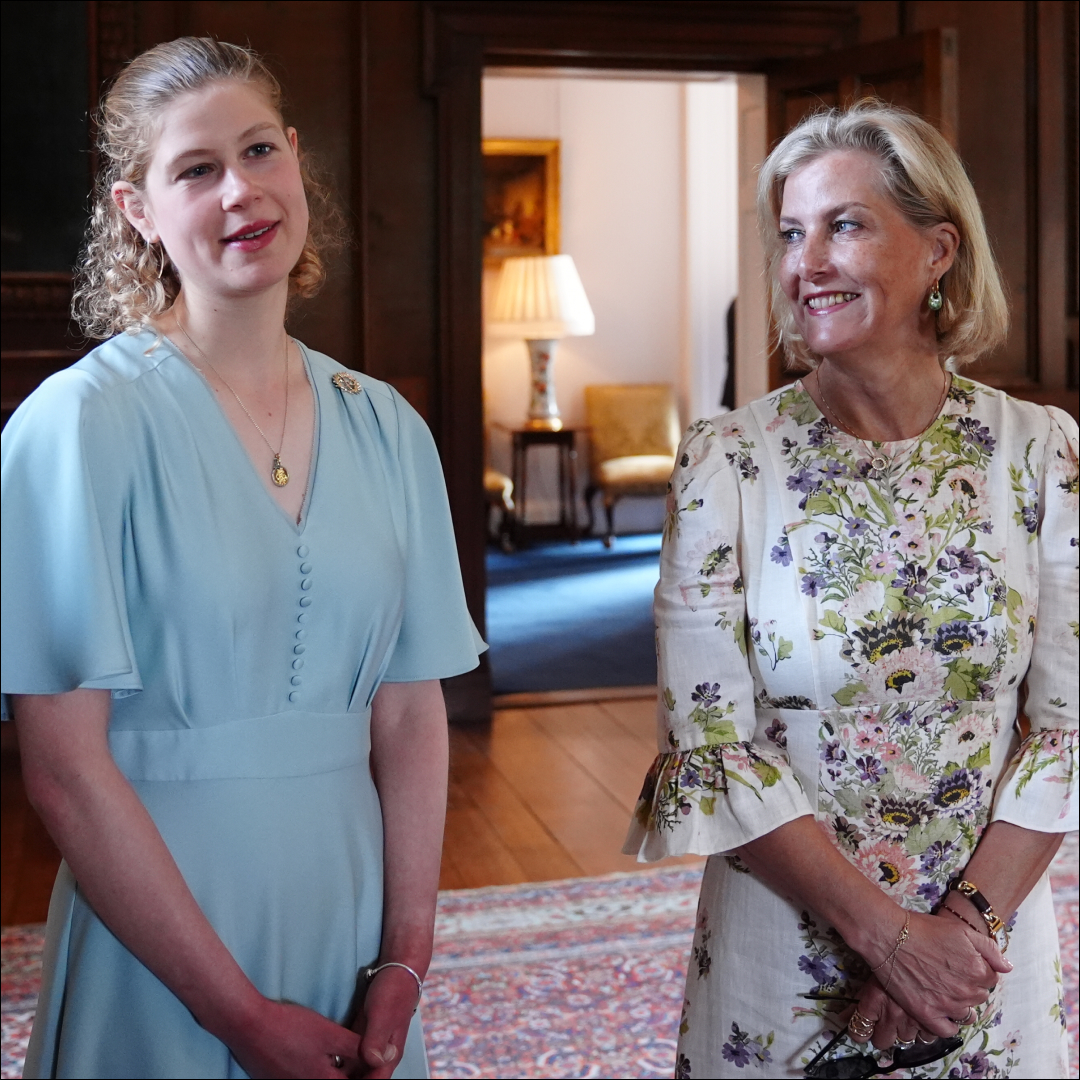 Lady Louise has blonde curly hair and is wearing a light blue dress while her mom Duchess Sophie has a blonde bob and is wearing a floral dress