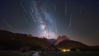 Perseid meteor shower where meteors streak through the star-studded sky above wooden alpine cabins. The band of the Milky Way stretches across the sky in the center of the image. 