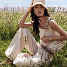 Young woman wearing Michael Kors white jeans, sweater bandeau, bag, and bucket hat sat in field.
