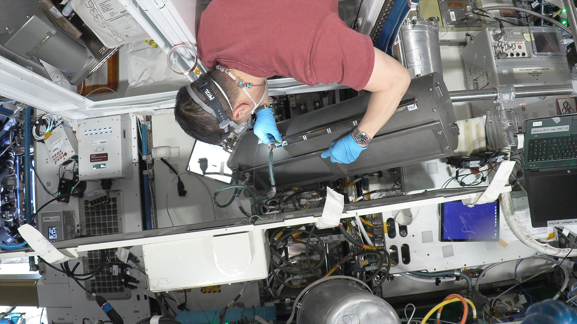 a man in a maroon shirt and safety goggles is seen from behind working to install a tank aboard a space station