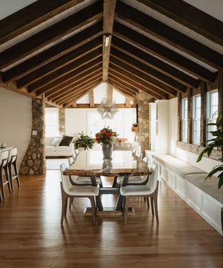 A farmhouse dining room with dark wooden beams in a triangular shape at the top, a wooden dining table with white leather seats and a vase of roses, dark brown wooden flooring, and stone pillars