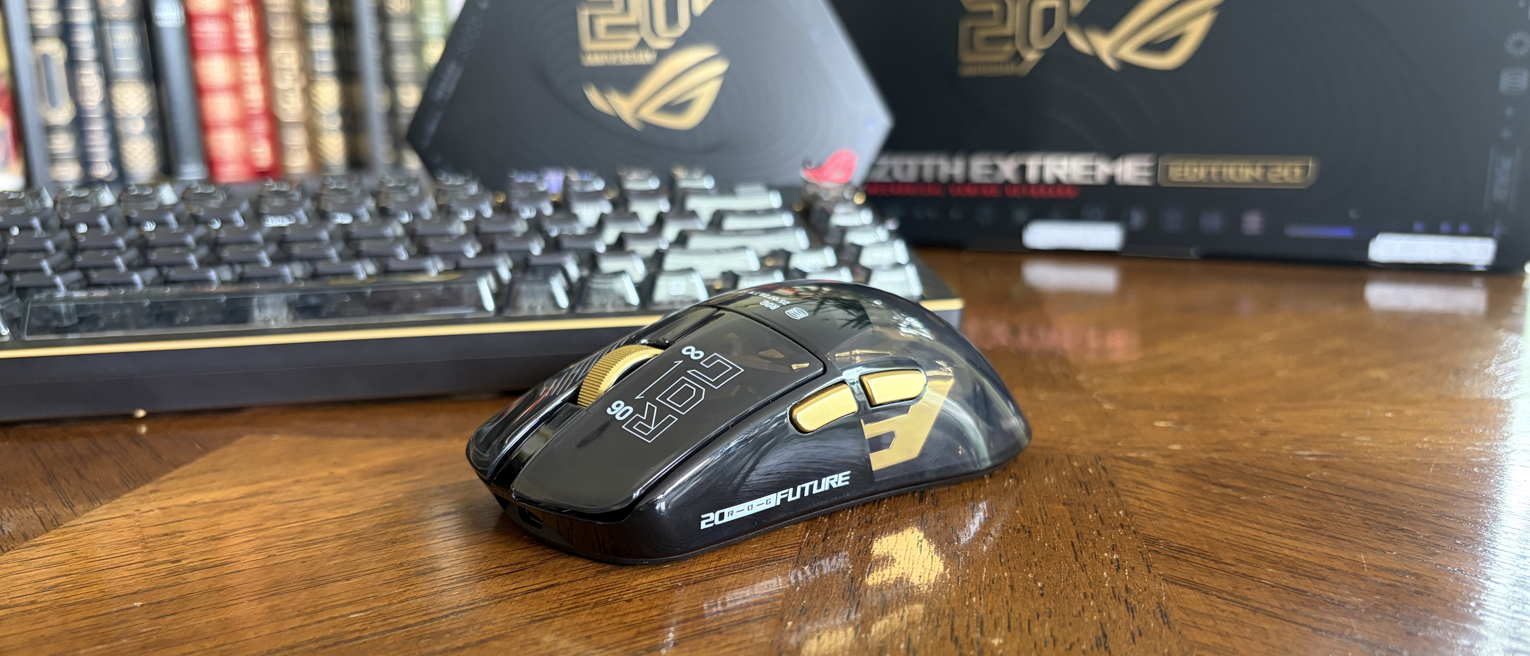 A black and gold ASUS ROG computer mouse on a wooden table, with a keyboard and bookcase in the background.
