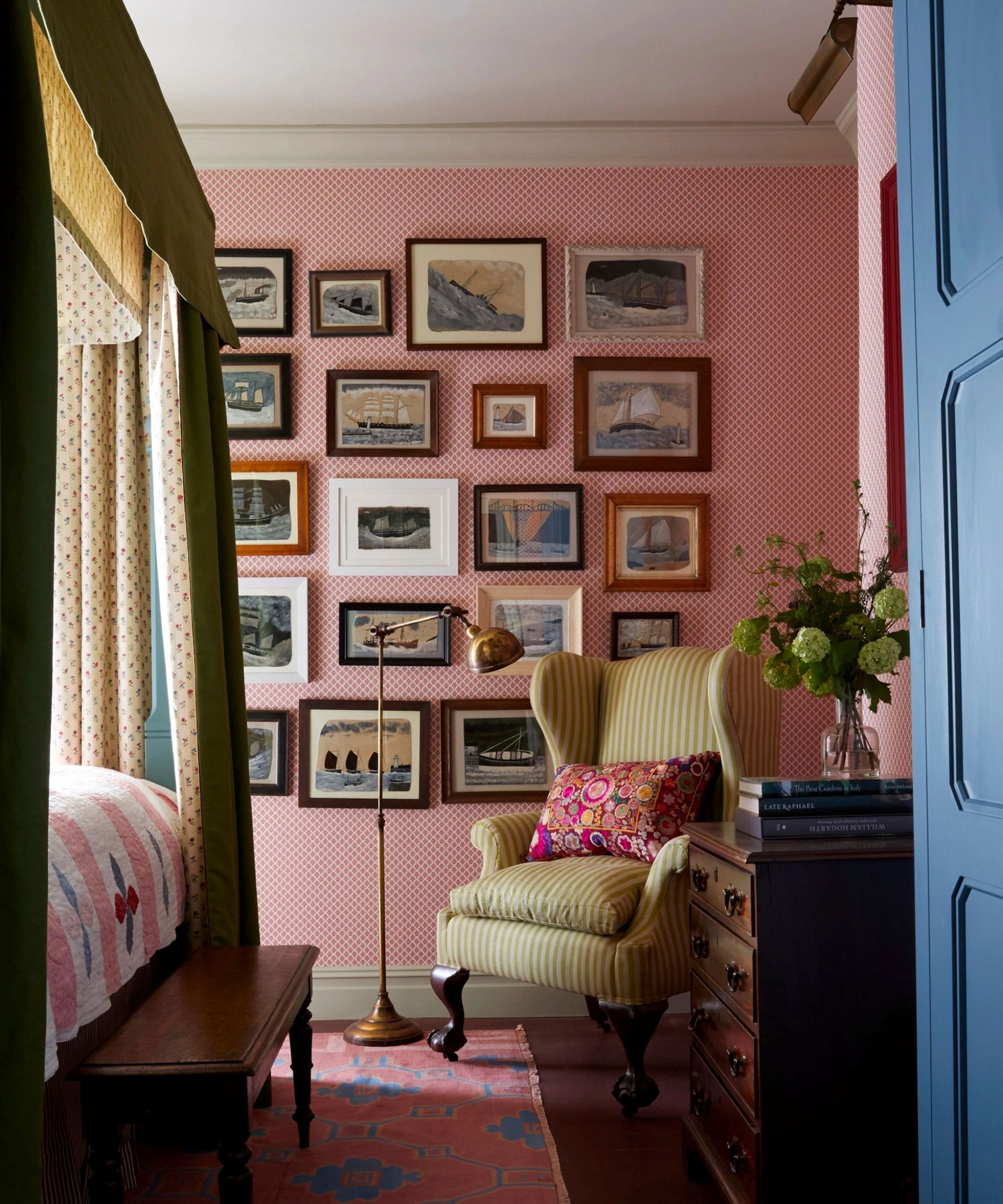 pink bedroom with a gallery wall of sea and boat paintings, a four posted canopy bed with mustard yellow and patterned drapes, a pink and blue rug, yellow armchair and floor lamp