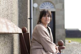 Shetland season 8 episode 5 - Stella Quinn (Dawn Steele) sits on a bench outside her family's house, with the front door behind her. She is wearing an oversized camel-coloured cardigan and jeans.
