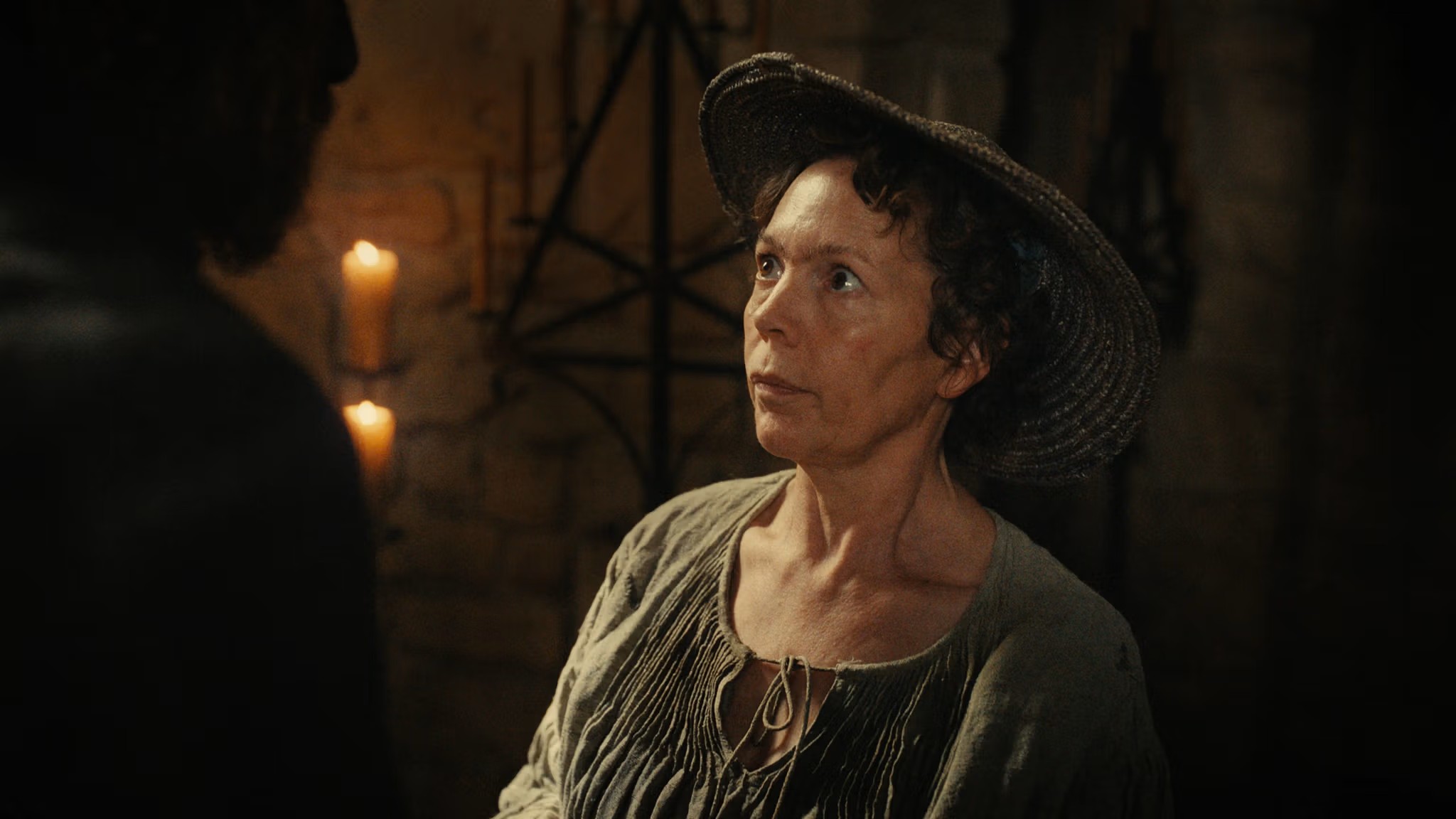 A middle-aged woman in a loose, green linen vest and a wide-brimmed wooden hat, standing in a candle-lit room while staring at a dark silhouette of a man in front of her.