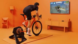 A person cycling on their bike in their orange living room cycling whilst their TV displays the Zwift virtual world