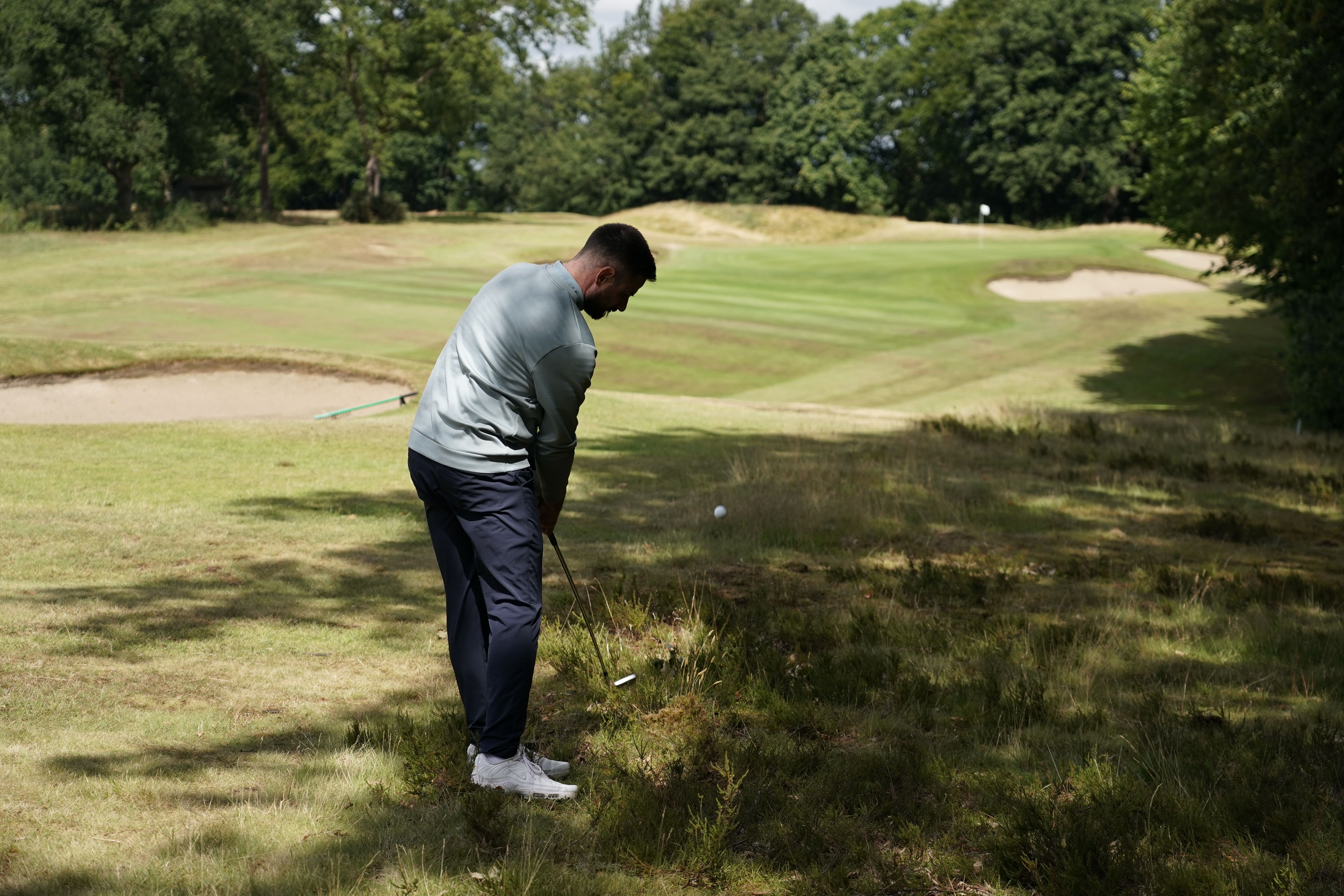 Baz Plummer hitting a golf shot in shadow cast by the trees to his right with a tough line into the green