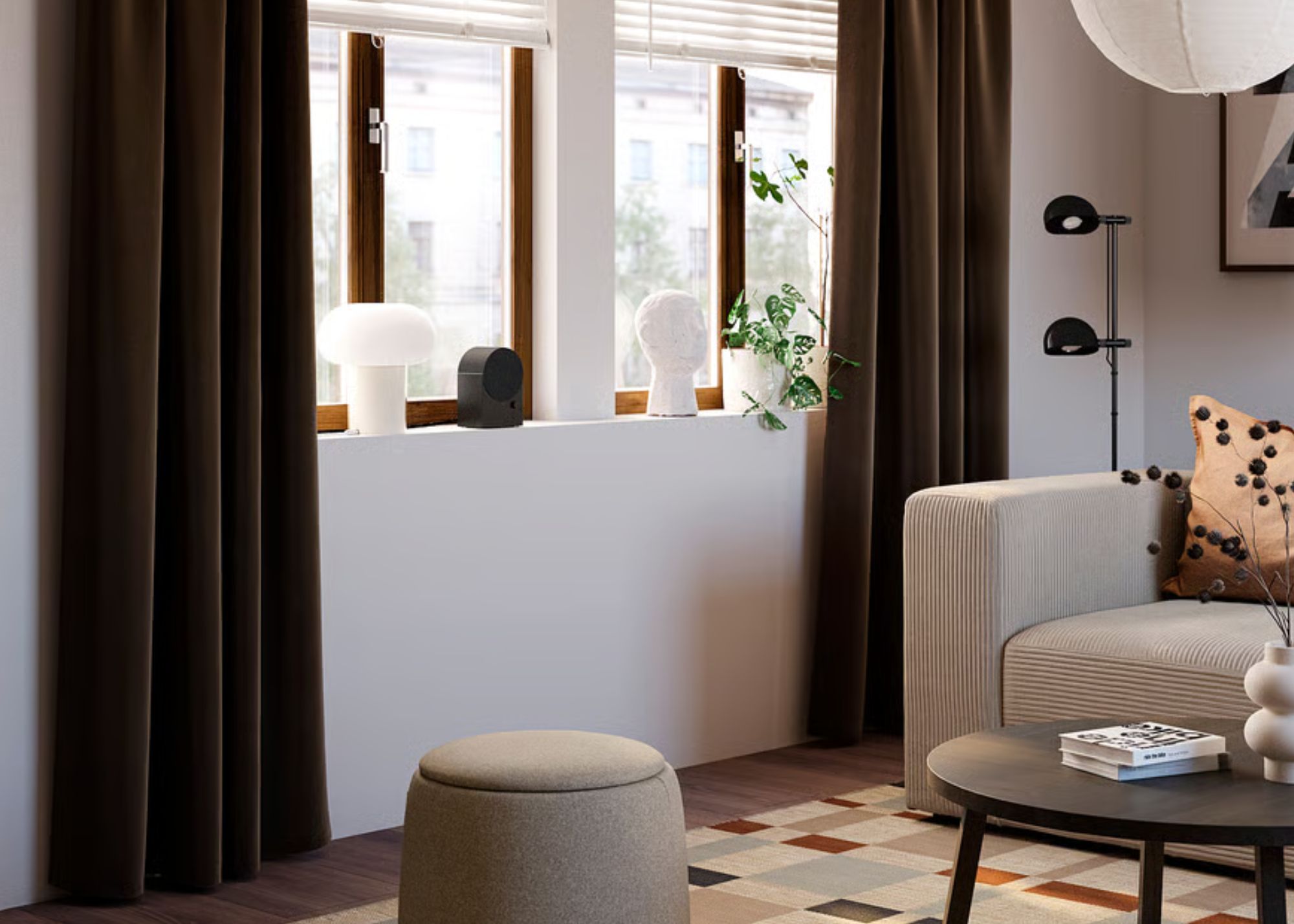 A modern living room with dark brown floor-length curtains and decor objects on the windowsill, a round dark wood coffee table and a beige sofa with a matching footstool