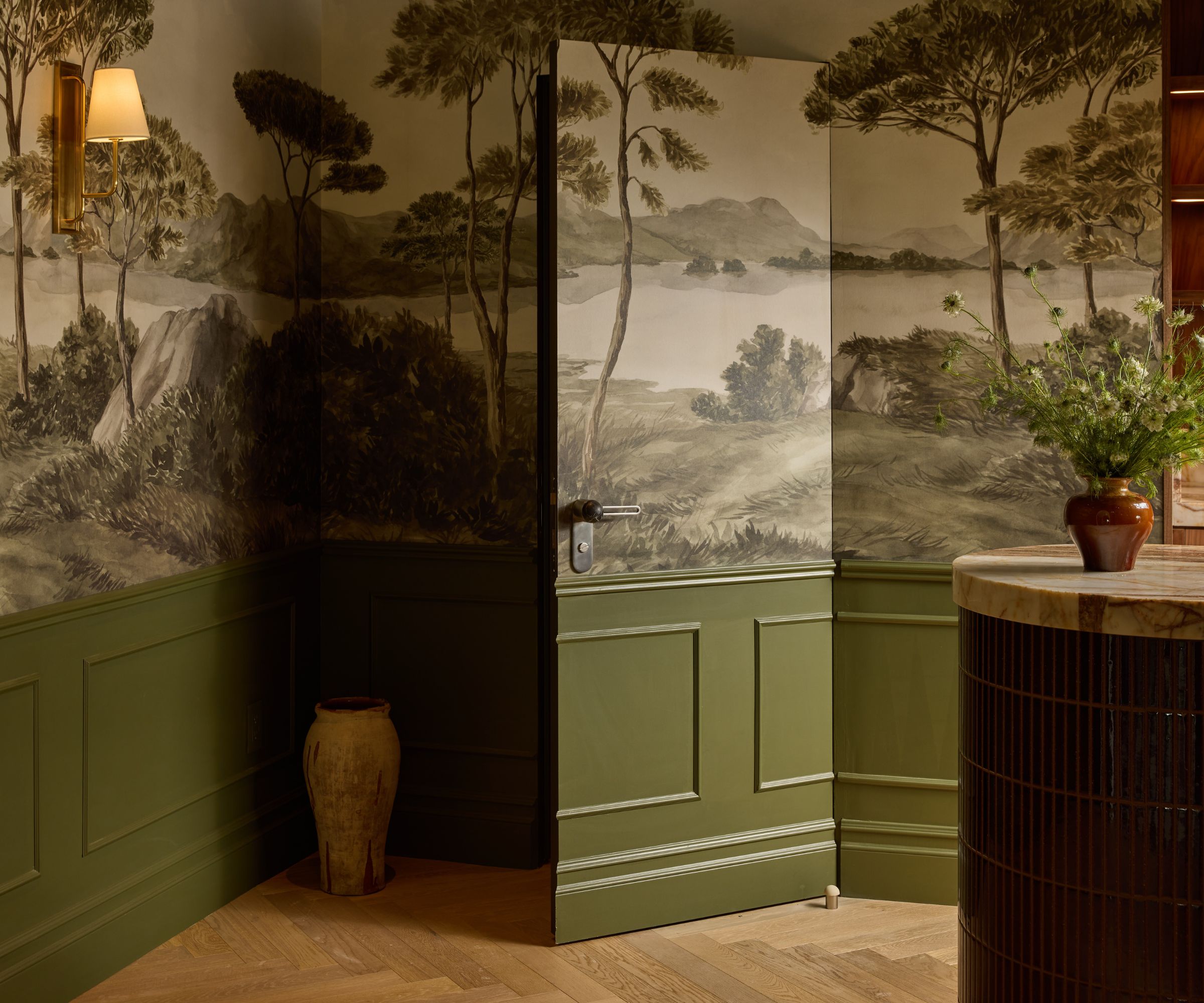 Close up of corner of room with green panelling and mural wallpaper across the top half of the wall, door into the room blends into the deisgn. Wooden flooring and marble countertop to right side.