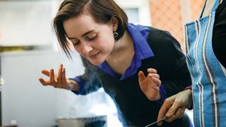 woman leans over a steaming pan on a stovetop and wafts its scent towards her face