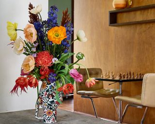 Colorful bouquet of flowers in patterned vase on table, wooden wall in background, small table with two chairs, chess set on table