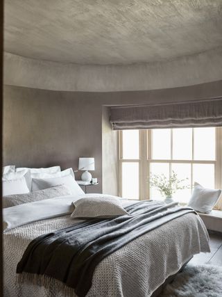 neutral bedroom with textured walls and ceiling, blinds, window seat, sheepskin rug, knitted throws, fringed blanket, white bedding, white table lamp, vase