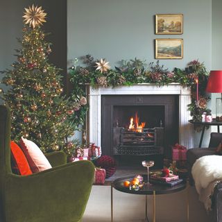 A sage green painted living room with a lit fireplace, a green velvet armchair and a decorated Christmas tree