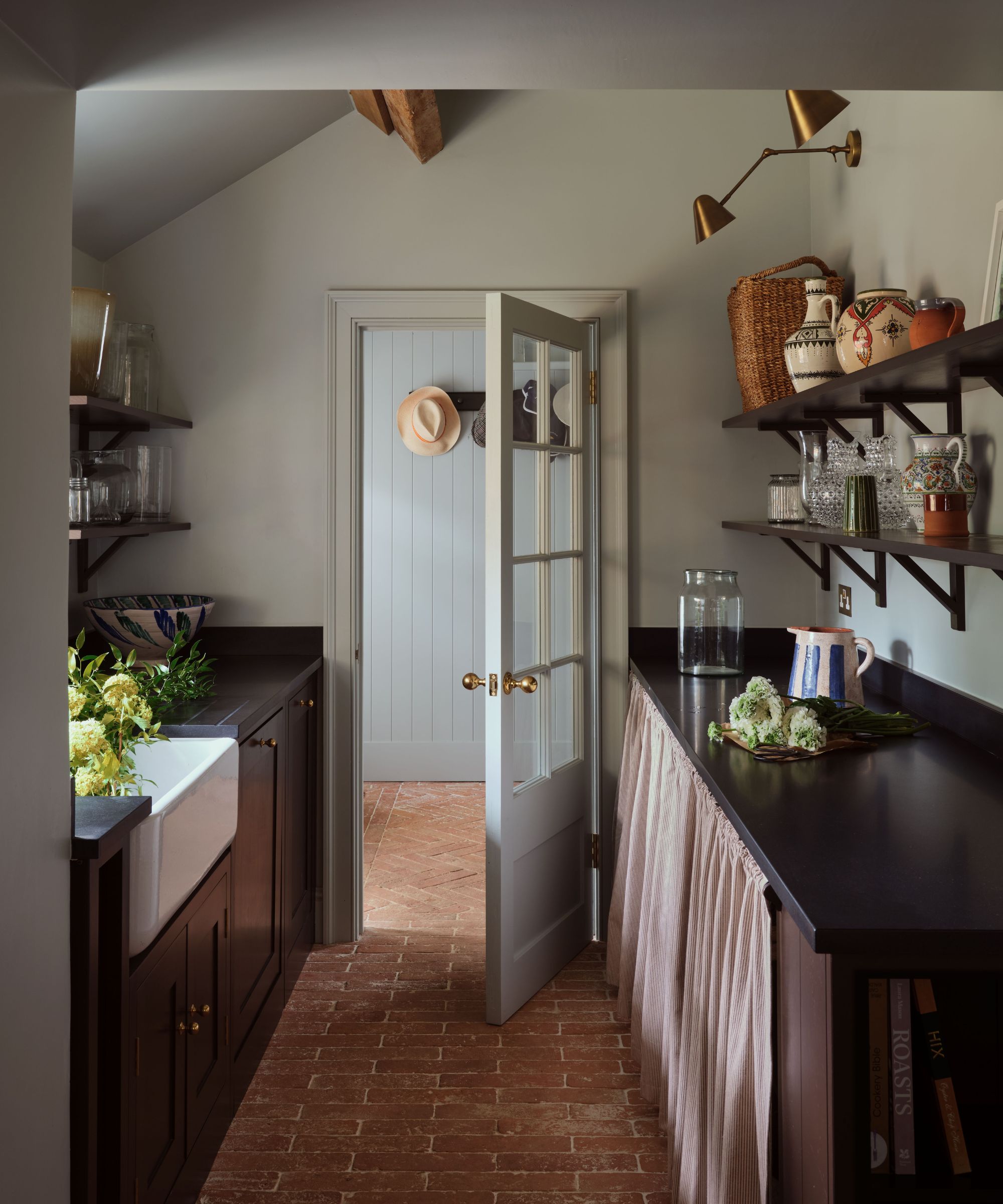 Pantry with shelves of vintage ceramics and glassware, black countertops with curtained storage and a white sink, gold sconces, pale blue painted walls, and exposed brick floor