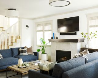 Bright living room painted white, stone fireplace with tv mounted on the wall above, twin blue sofas facing one another, central low leather coffee table, decorated with plants, flowers and cushions