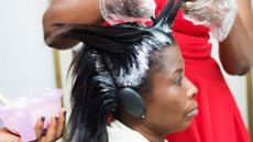Close-up image of a Black woman receiving a hair-straightening treatment in a salon. A hairdresser (whose face cannot be seen) wearing a red dress and gloves can be seen applying the white cream to the woman's hair while another person's hand can be seen holding a plastic tub containing the cream.