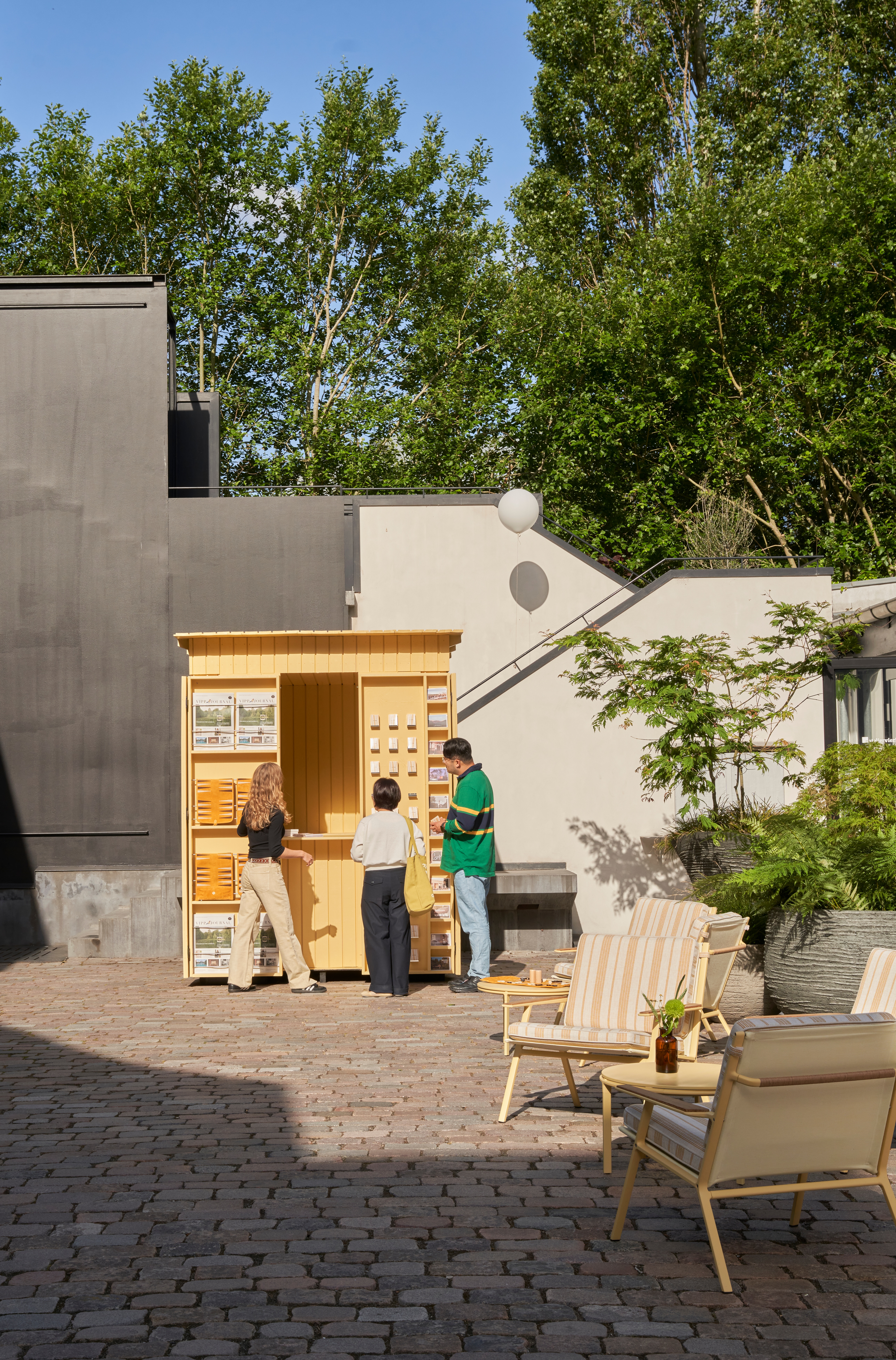 A playful installation comprising a chrome-clad, two-seat rocking chair, a butter yellow-painted kiosk, and a massive modular sofa with a square surface, butter yellow fabric, and chrome detailing.