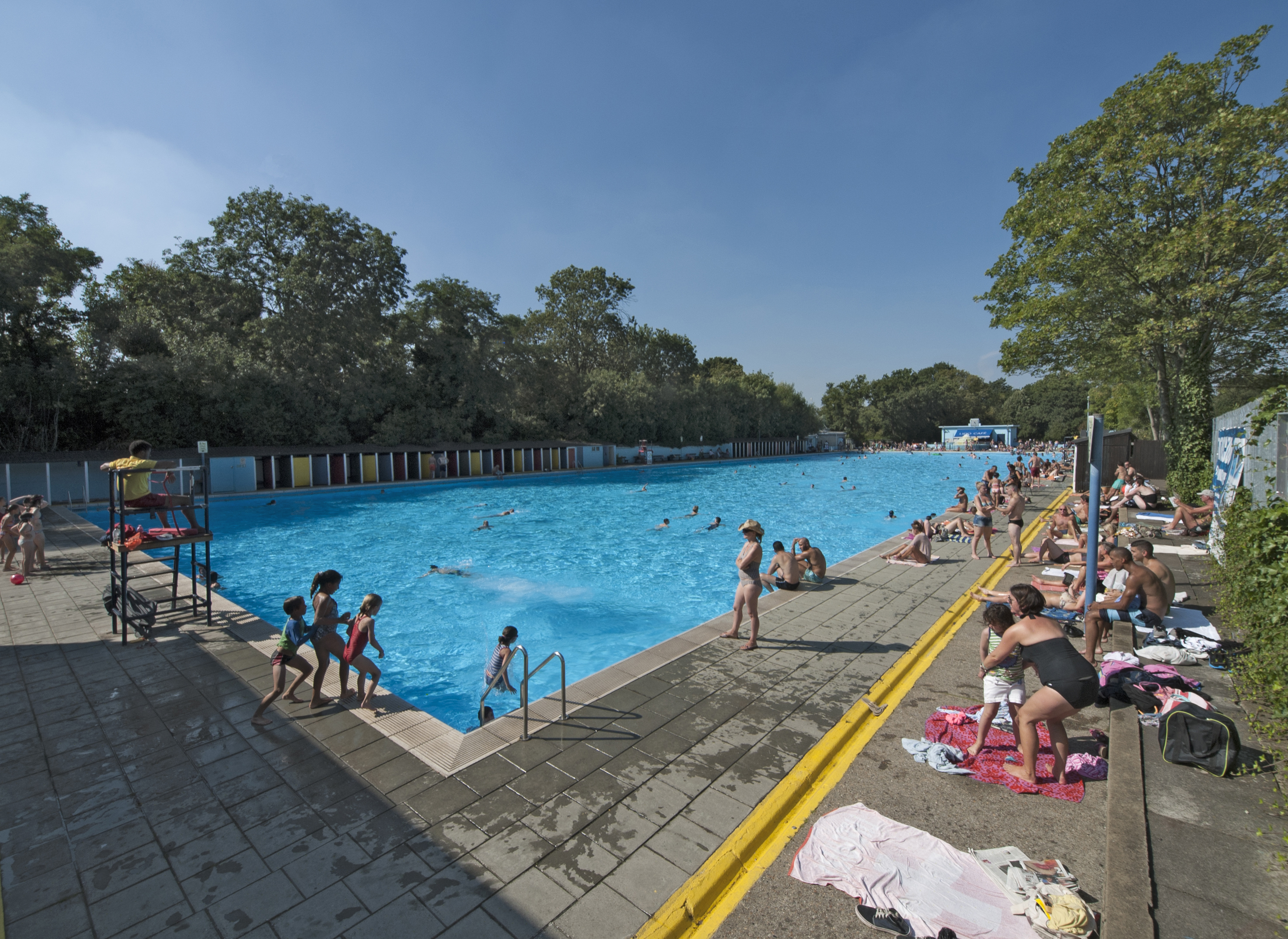 an elevated view of the swimming pool at Tooting Bec Lido, Streatham, busy with swimmers and sunbathers, seen from the south-east corner. Tooting Bec Lido is London's largest and oldest outdoor pool.
