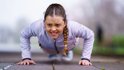 Woman with look of determination on her face midway through a push-up. She is wearing a sports jacket and is outside
