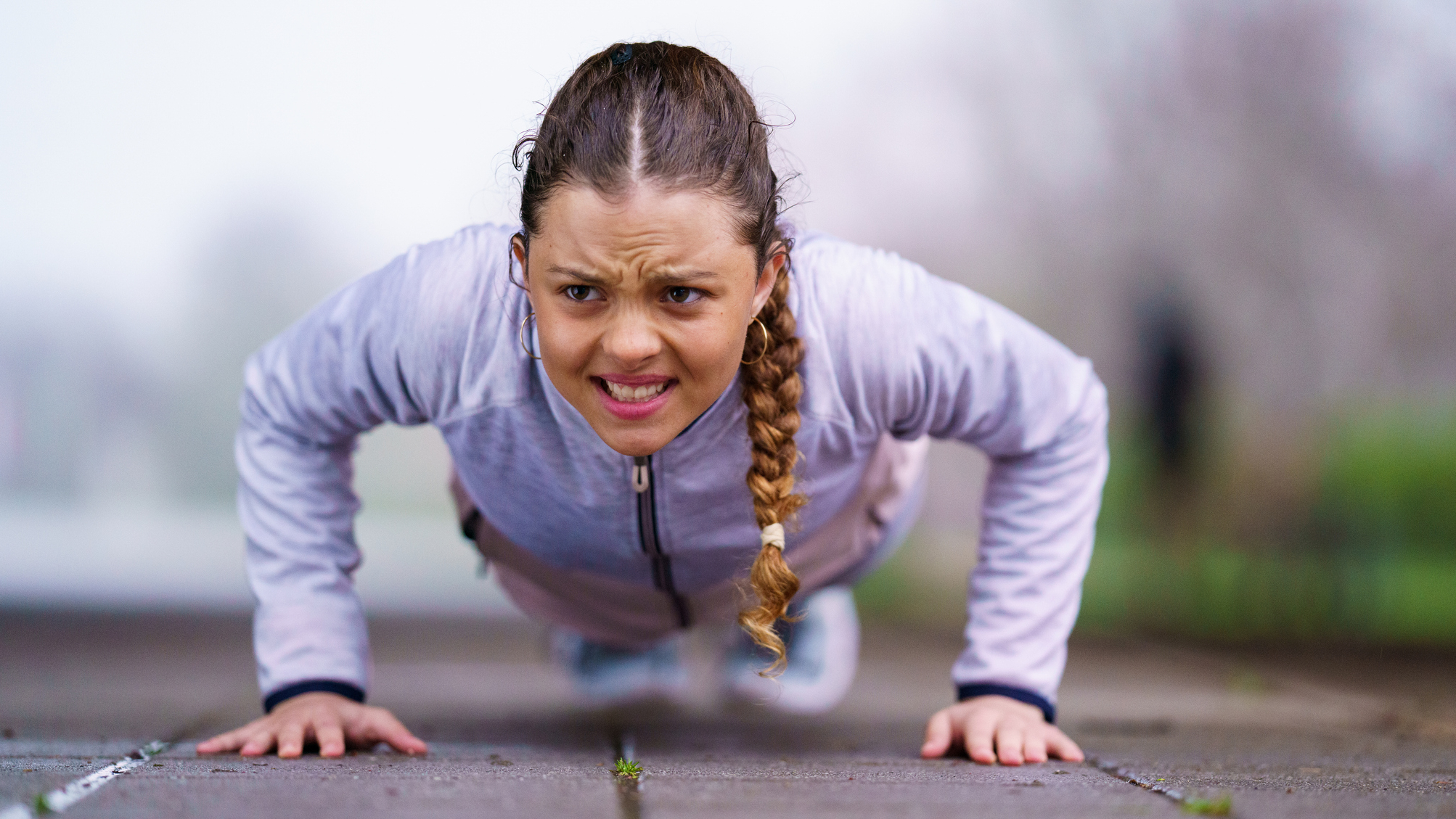 Woman with look of determination on her face midway through a push-up. She is wearing a sports jacket and is outside