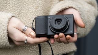 The Ricoh GR IIIx being held in a person's hands wearing a beige jumper