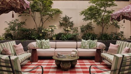 Garden with plants growing on the wall, a large red gingham rug with a large cream sofa on top, green metal armchairs with striped green cushions, and a marble green round coffee table at the centre.