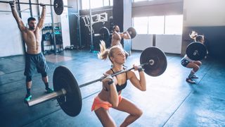 Four people perform thruster exercise with barbells in a gym