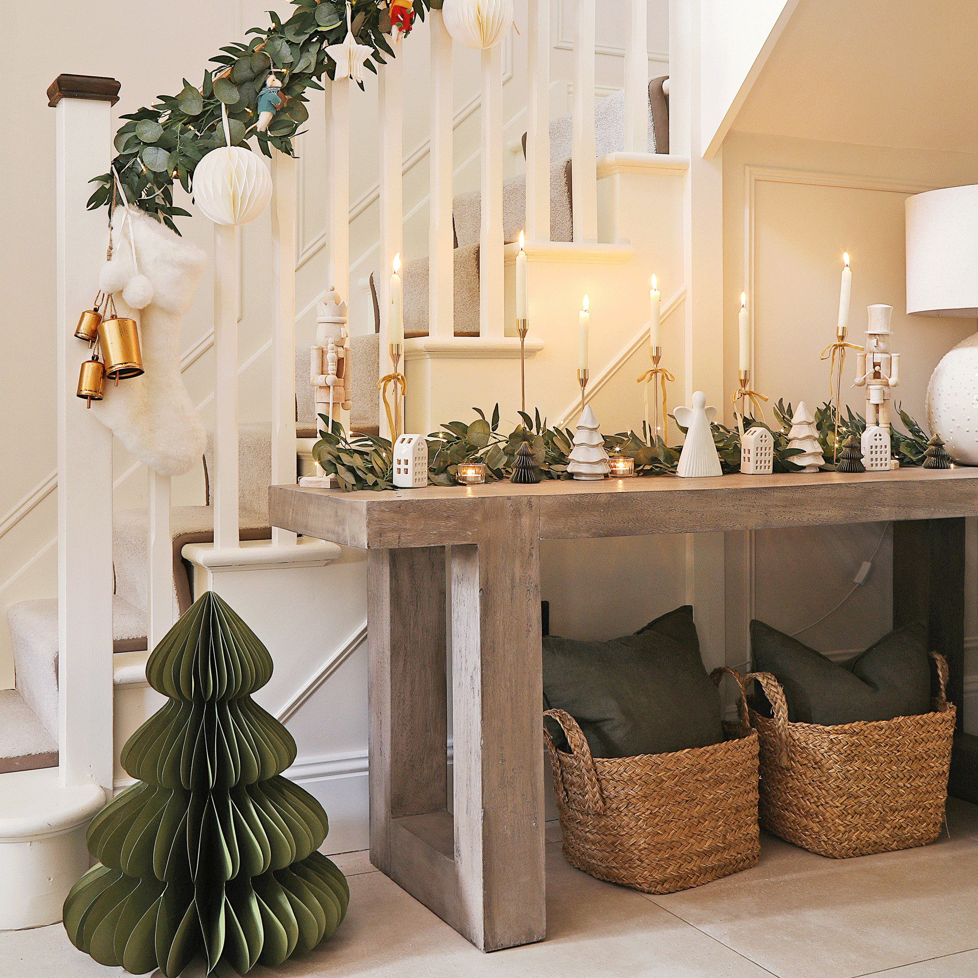 a hallway and staircase with a paper Christmas tree, garland on the handrail, console table filled with Christmas decorations and candles