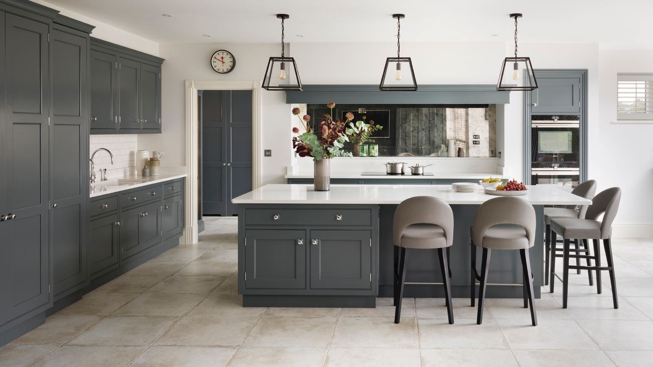 Classic style kitchen with white countertops, gray cabinets, pale gray tiles, bar stools, metal and glass pendant lights, mirror, vase of flowers,