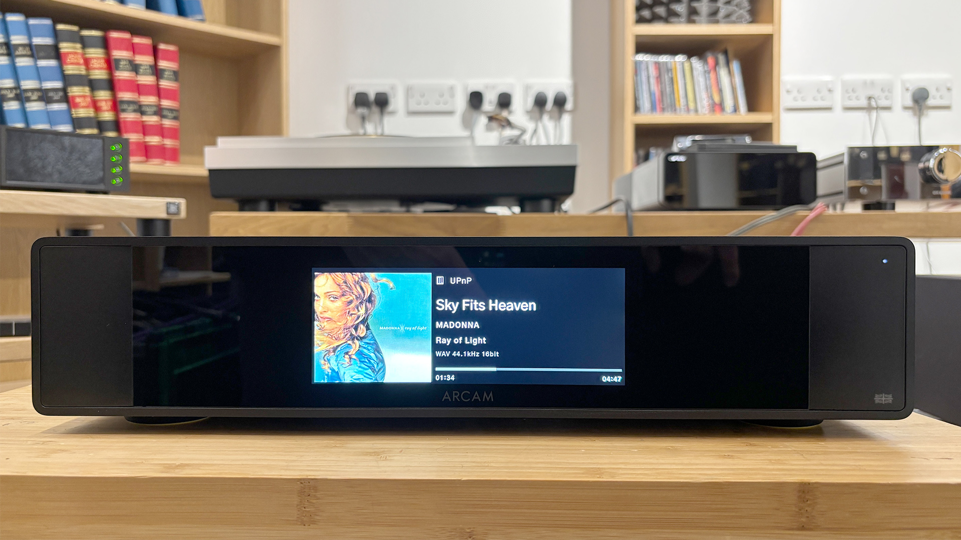 Arcam ST25 music streamer on wooden AV rack in busy test room, display shows Madonna