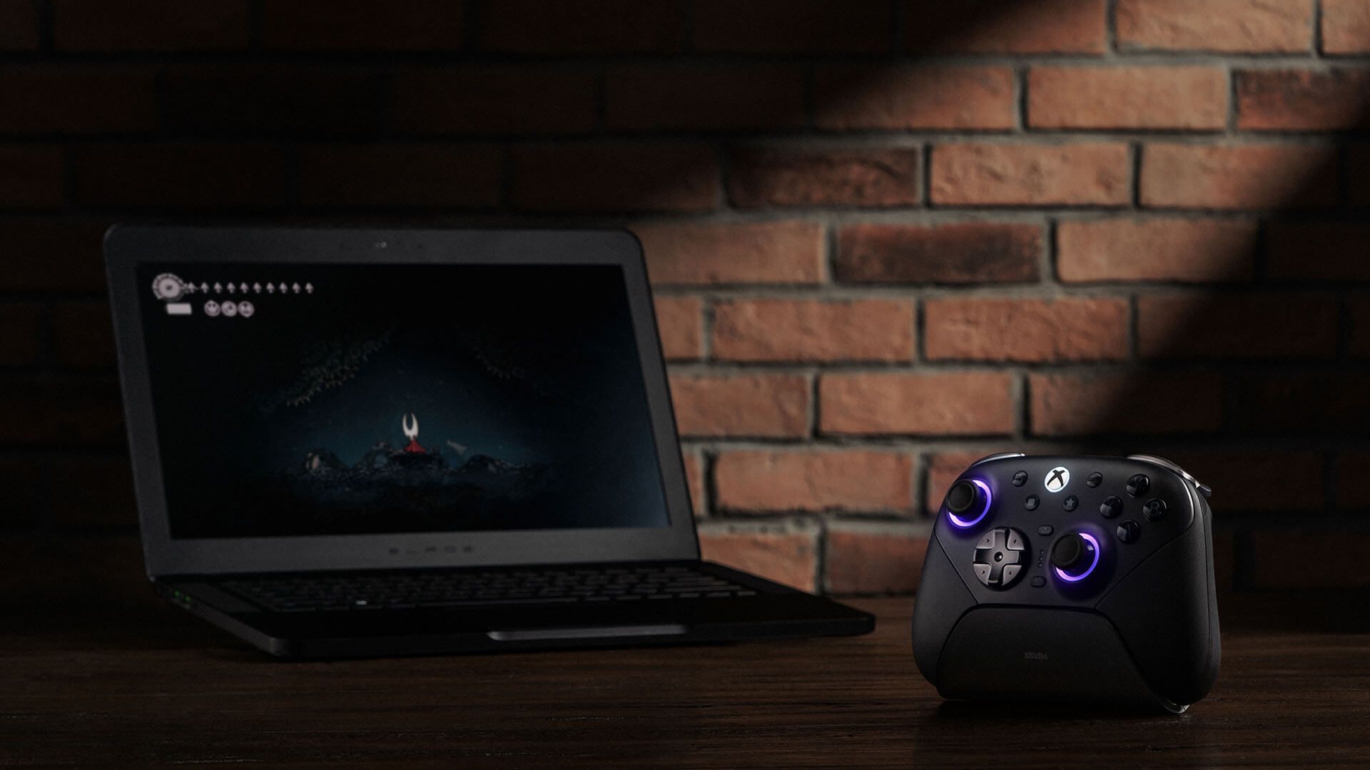 An 8BitDo Ultimate 3E controller next to a gaming laptop that's playing Hollow Knight