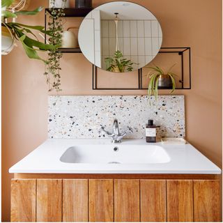 white basin with wooden drawer underneath, earthy wall colour, circular mirror and green plants on black brackets