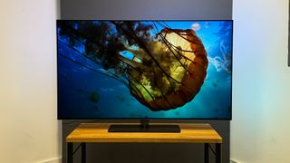 The Philips OLED808 on a wooden stand in front or a white and grey background. A very large jellyfish is on the screen.
