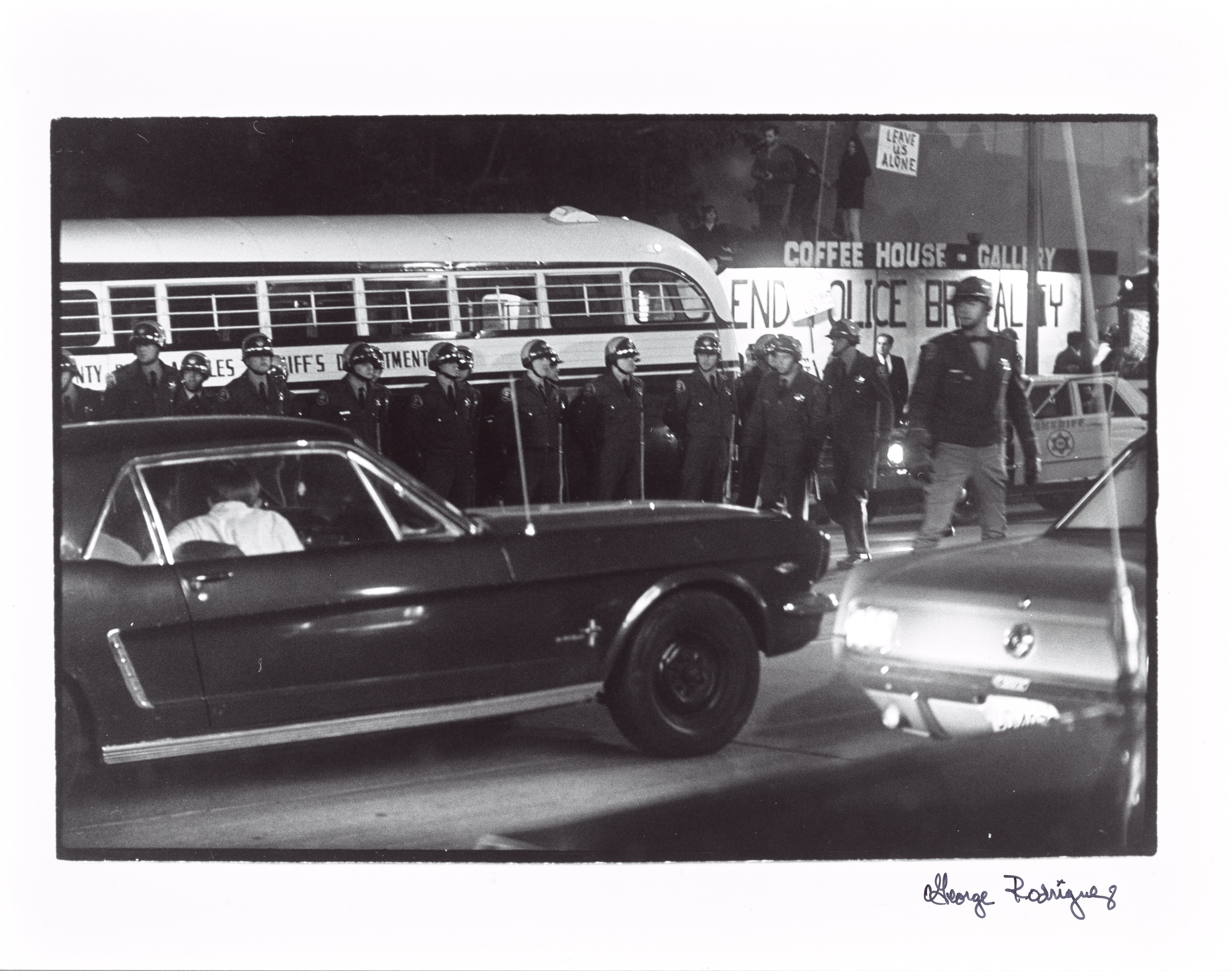 Black and white photograph of a row of helmeted police officers standing behind cars on a night-time street, with a "END POLICE BRUTALITY" sign visible on a bus in the background.