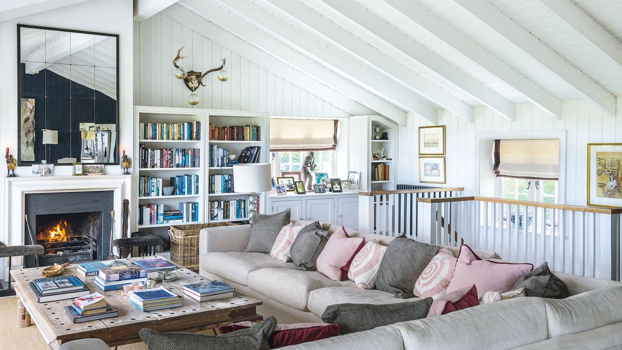 living room with beige corner sofa and pink cushions with fire lit and stairs behind sofa and white wooden cladding walls and ceiling