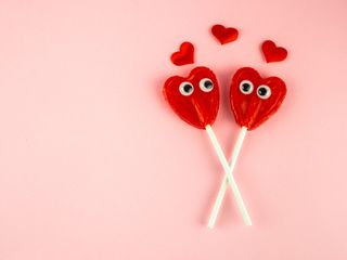 A light pink background with two red heart-shaped lolly pops with eyes together with small red hearts.