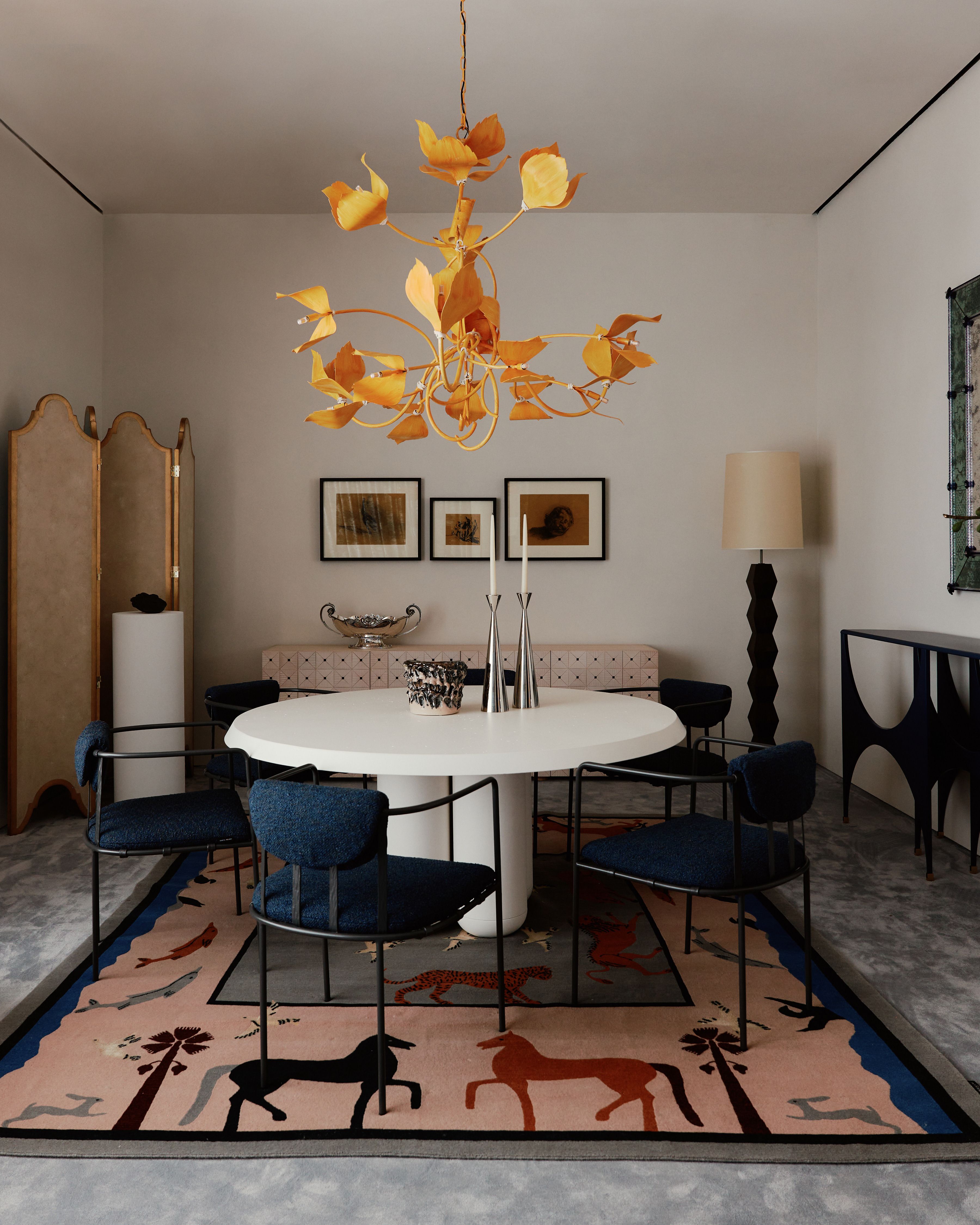 A warm, inventively decorated showroom features a rose-tinted rug with stylized animal motifs, upholstered chairs in blue and black, framed artwork, and stunning wooden carbinetry.
