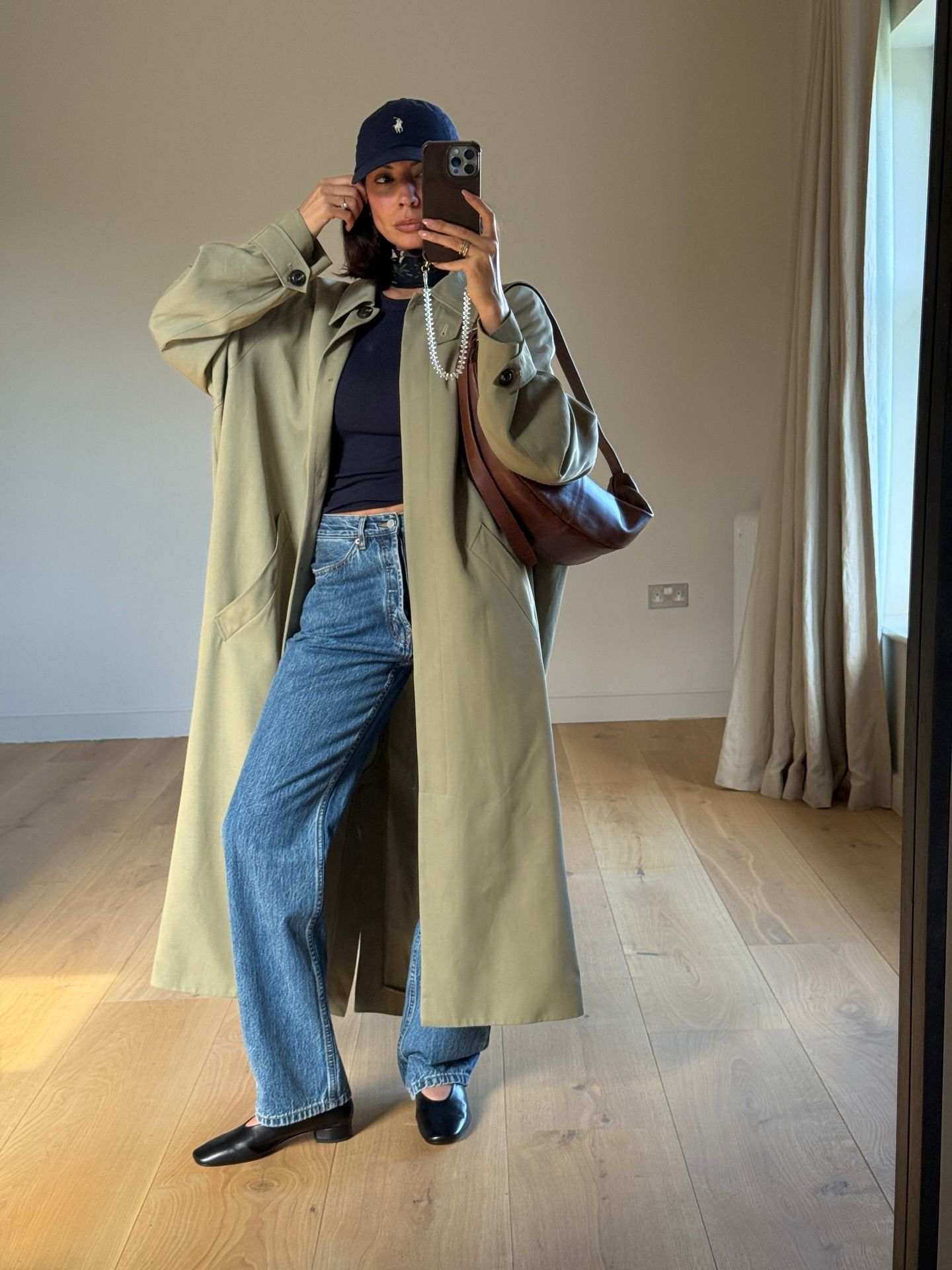 Influencer takes a mirror selfie wearing jeans, heels high vamp shoes and a khaki trench coat.