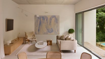 A minimalist living room with a framed TV, a wood sideboard, rattan chairs, a woolly sofa chair, a linen sofa, and a coffee table, by a large painting