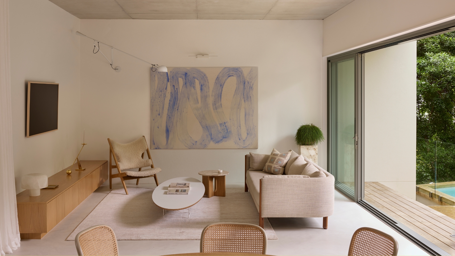 A minimalist living room with a framed TV, a wood sideboard, rattan chairs, a woolly sofa chair, a linen sofa, and a coffee table, by a large painting